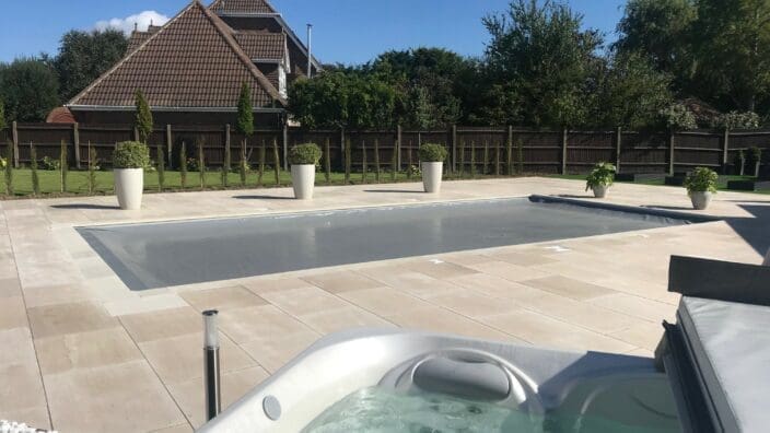A covered rectangular pool sits on a pale tiled patio; a bubbling hot tub fills the foreground, with potted shrubs, a wooden fence and a house with tiled roofs beyond.