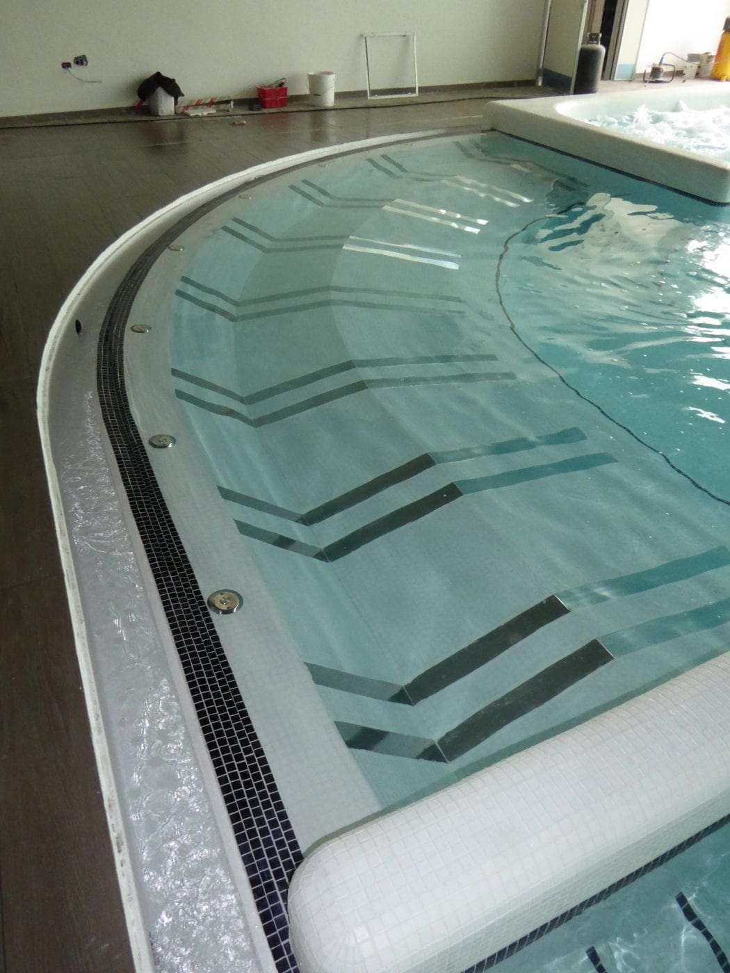 Curved indoor pool steps, water rippling over tiled risers with chevron stripes while an adjacent raised spa bubbles, tiled overflow channel and dark wood deck with tools against far wall