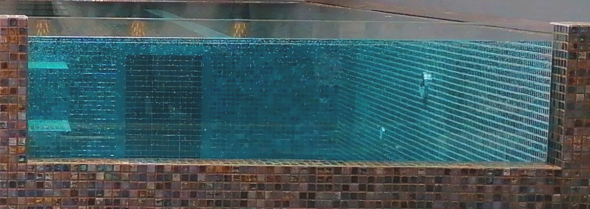 Glass-sided indoor pool displays clear turquoise water rippling against a tiled interior; mosaic brown exterior cladding, submerged corner steps to left, small wall lights casting reflections across the surface.