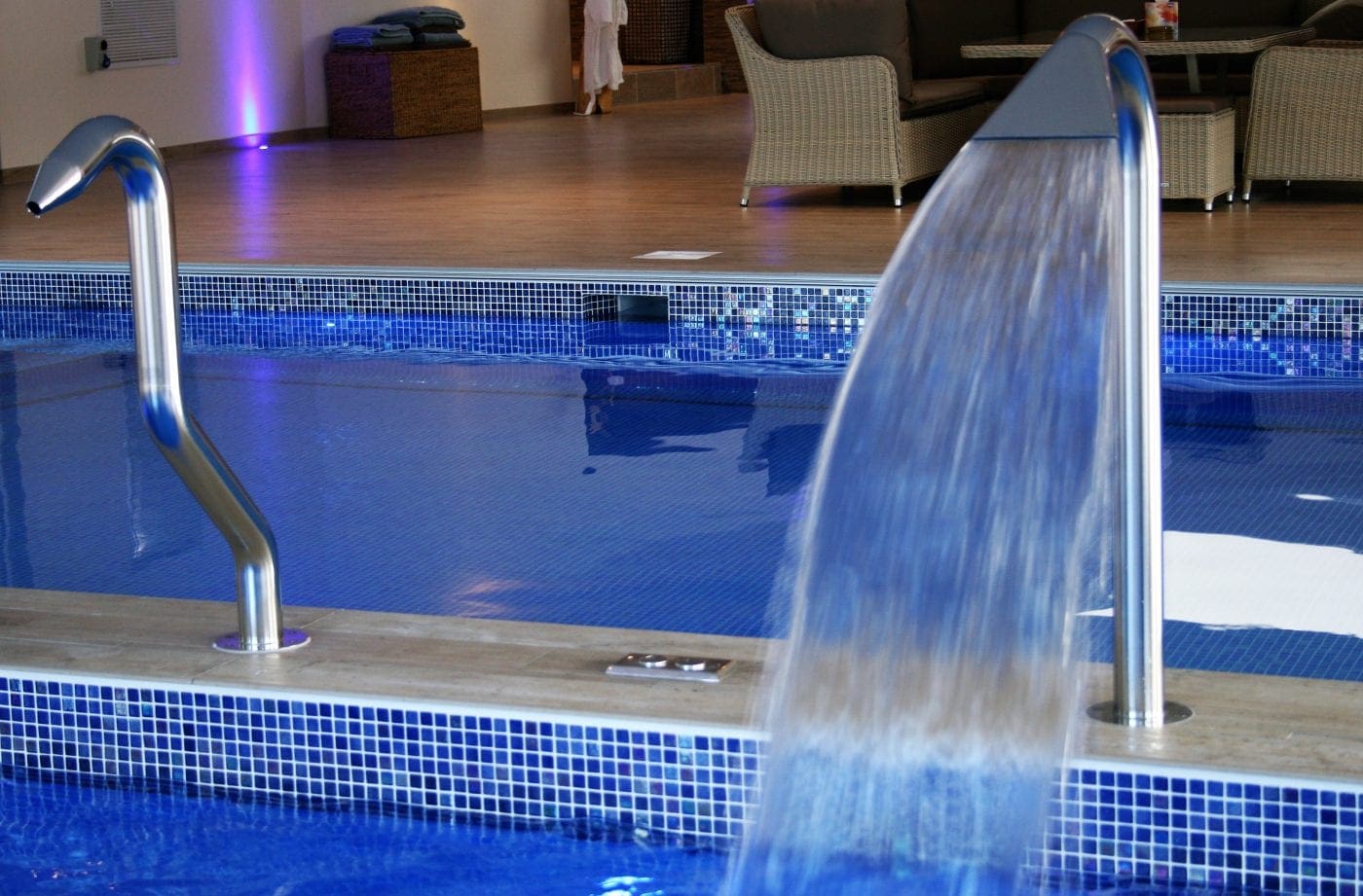 Curved stainless steel spout pours a wide sheet of water into a tiled indoor pool, a second handrail beside it, with wood decking and wicker lounge chairs in the background.