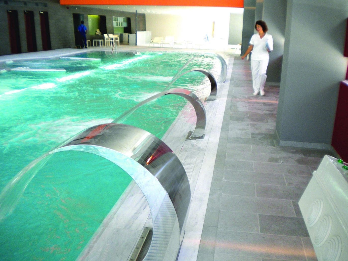 Metallic arched fountains spout curved streams into a turquoise indoor pool, while a woman in white uniform walks along the tiled poolside amid lounge chairs and a modern spa-like interior.