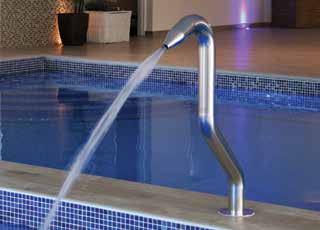 Stainless-steel curved water spout arcs a clear stream into a blue-tiled indoor swimming pool, set against a tiled deck with potted plants and soft ambient lighting.