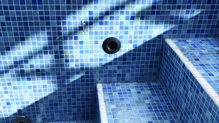 Blue mosaic-tiled pool steps sit empty, sunlight casts diagonal shadows across the tiled wall; a circular drain opening and a small brown leaf lie on the steps in the pool.