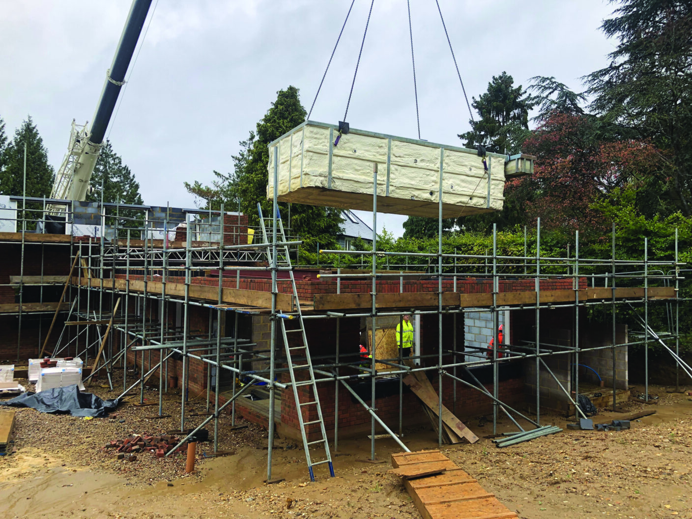 A large rectangular insulated module is being hoisted by a crane over metal scaffolding around a partially built brick house on a muddy lot with workers and trees.