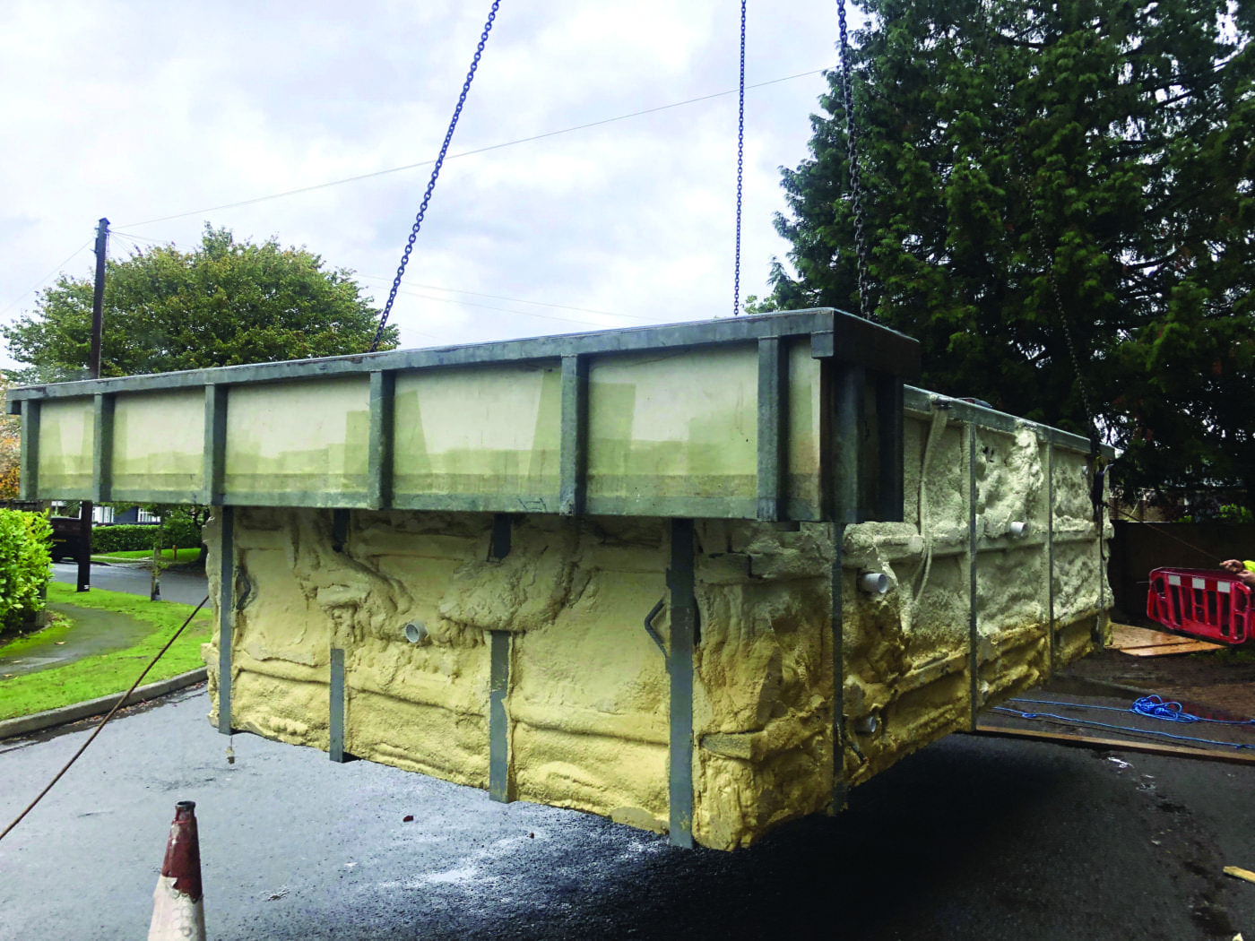 A large rectangular fibreglass pool shell suspended by chains being lifted above a wet residential street, foam-insulated sides and metal frame visible, traffic cone and trees in the background.