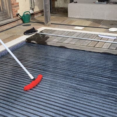 Pool cover being brushed with a red broom on a residential patio beside stone tiles and a wooden deck, near a green watering can and house steps.