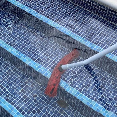 A red pool vacuum head attached to a white hose scrubs water along tiled blue mosaic steps while small ripples and light reflections distort the pool surface.
