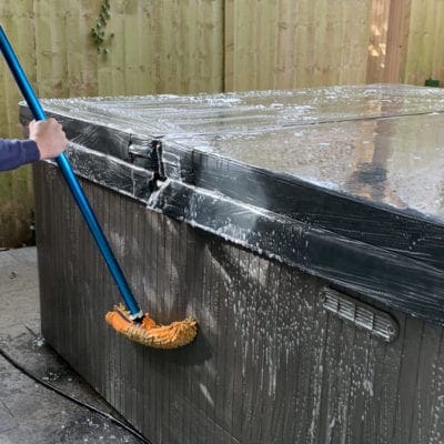 A rectangular hot tub is being scrubbed with a soapy orange brush on a blue pole; suds and water streak its gray sides on a patio by a wooden fence.