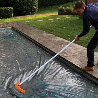 Long handled orange brush is pushed across a water covered pool cover by a man standing on the stone edge of pool in a trimmed backyard with hedges and grass