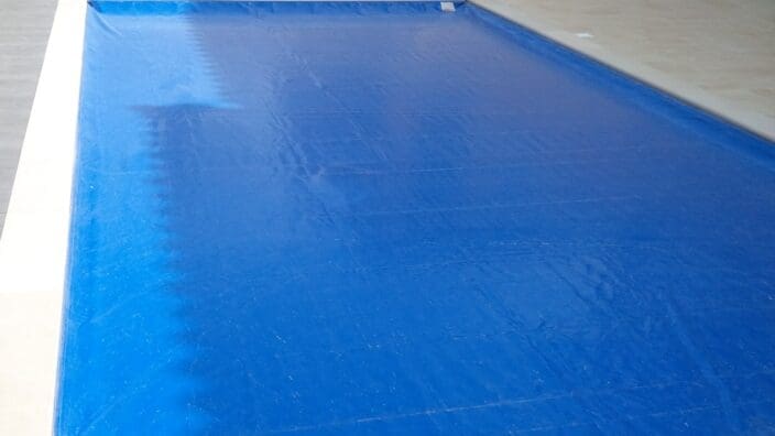 Rectangular swimming pool covered by a taut blue safety tarp, sitting in a tiled backyard patio with striped outdoor sofas, potted plants, and a lawn border in sunlight.