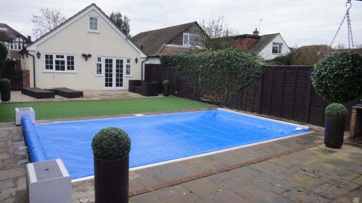 Rectangular in-ground pool is covered by a blue tarp in a paved suburban backyard with a cream bungalow, patio furniture, potted spherical topiary and an ivy-covered dark fence © www.funfantastic.com