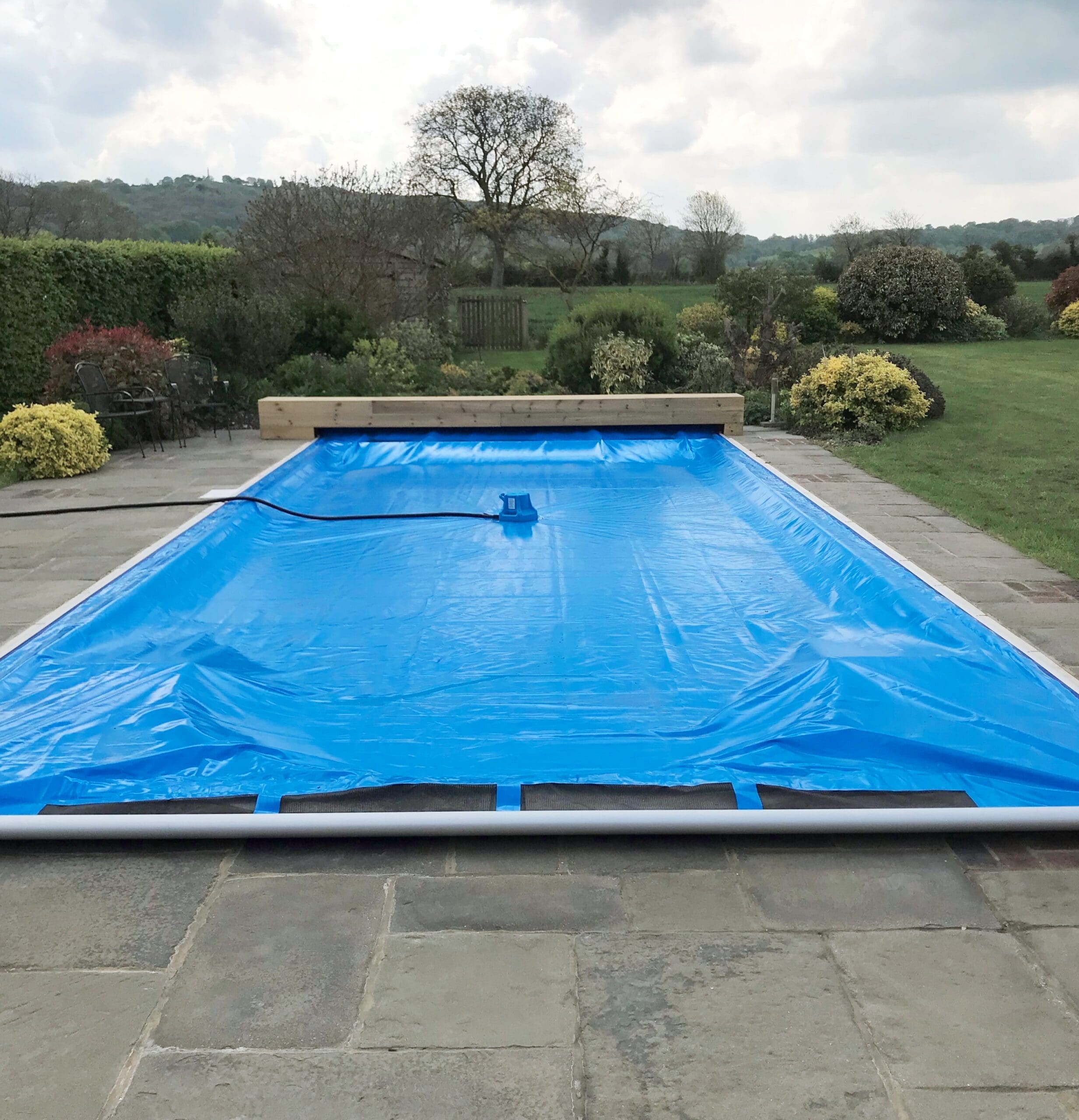 Rectangular outdoor swimming pool covered by a blue solar cover with a small pump and hose resting on it, set on a stone patio surrounded by shrubs, lawn, trees and distant hills.