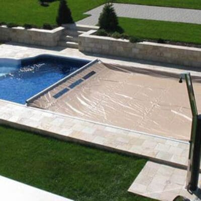 Rectangular in-ground swimming pool with rounded spa, one half filled with blue water and the other half covered by a beige cover, set in stone patio surrounded by green lawn.