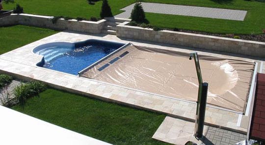 Rectangular in-ground swimming pool with rounded spa, one half filled with blue water and the other half covered by a beige cover, set in stone patio surrounded by green lawn.