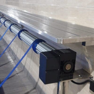 Stainless-steel bench shelf mounted on a tiled wall, supported by a tubular roller underneath with blue straps wound around it; a black motorized end cap and support leg.