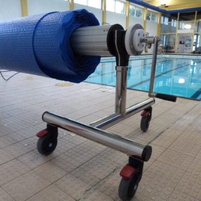 Pool cover roller is mounted on a wheeled stainless-steel frame, holding a blue solar pool cover partially rolled, positioned beside an indoor swimming pool on tiled deck under windows.