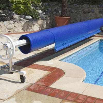 Blue solar pool cover is rolled on a metal reel, partially extended over water, positioned on a tiled deck with brick coping beside an in-ground pool and stone wall planter.