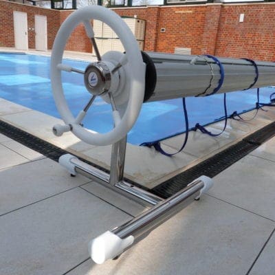 Pool cover roller sits at pool edge, white handwheel turning an aluminum tube winding a blue cover; tiled deck and brick wall enclosing the outdoor swimming pool.