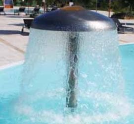 Mushroom-shaped stainless-steel fountain shedding a continuous curtain of water from its domed top onto a shallow light-blue pool, surrounded by paved deck and benches in an outdoor plaza.