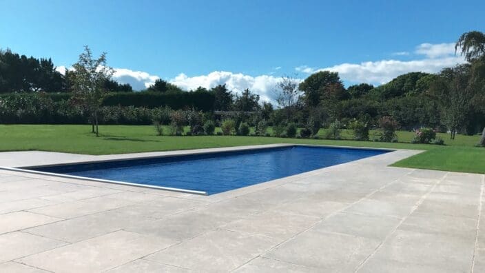 Rectangular in-ground swimming pool sits covered with a taut blue safety cover, recessed in a pale stone patio, surrounded by trimmed lawn, scattered trees and hedges beneath a blue sky.