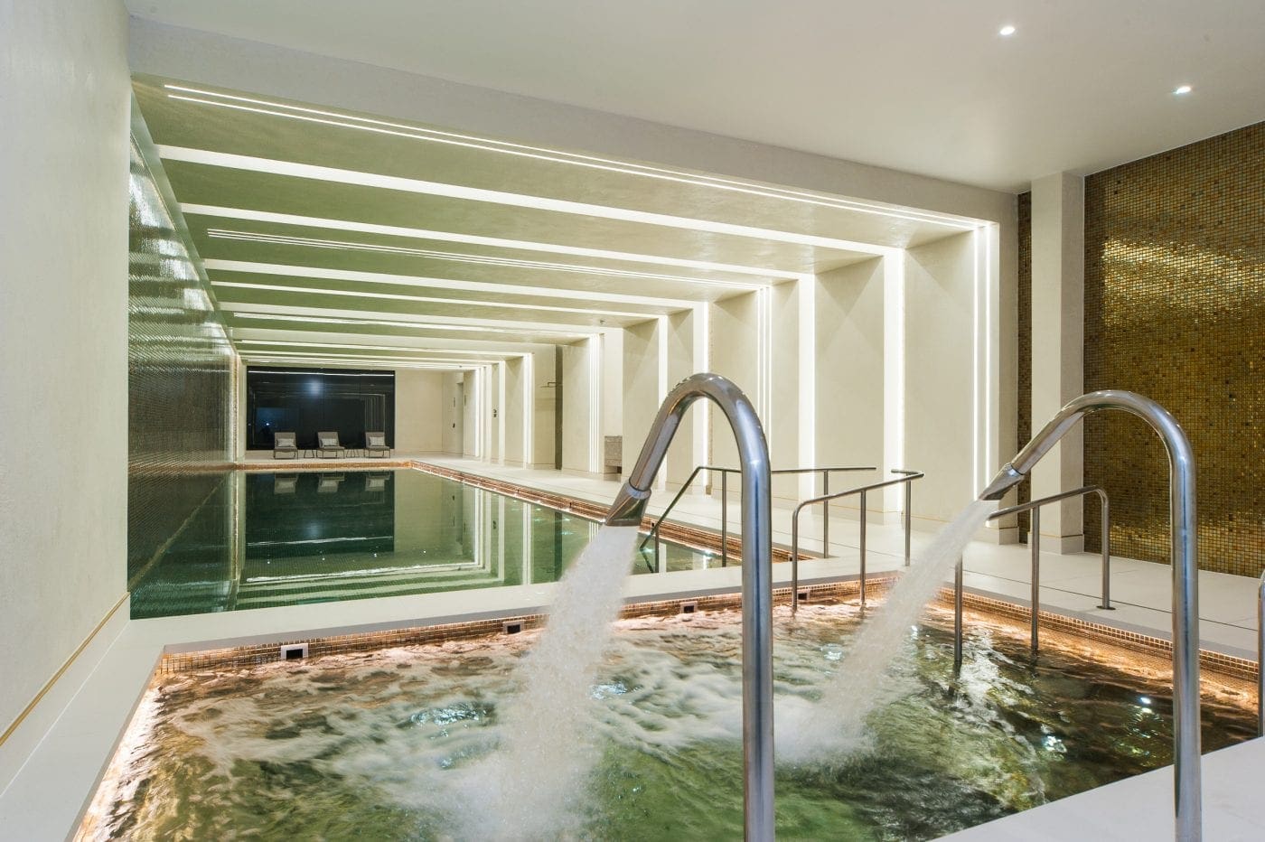 Stainless-steel spa jets pour water into a bubbling tub; a long lap pool stretches behind with lounge chairs; tiled room lit by linear ceiling lights and a gold mosaic wall.