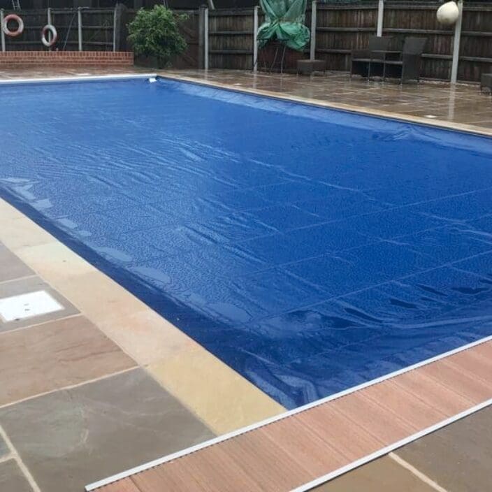 Rectangular outdoor swimming pool covered by a blue solar cover, sitting on a wet stone terrace with metal ladder, life rings and wooden fence in the background.
