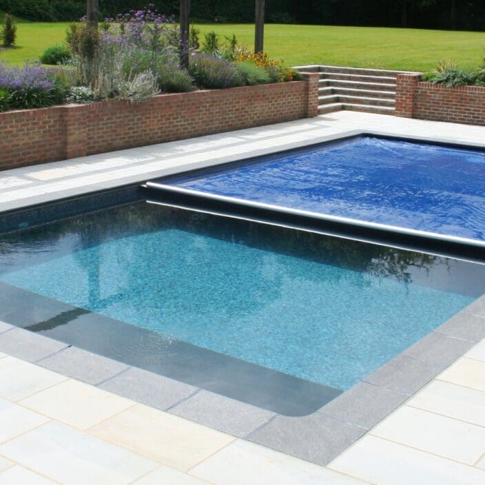 Rectangular swimming pool partially covered by a blue solar cover, reflecting sunlight and gentle ripples; set in pale tiled patio with brick retaining wall, flowerbeds and a manicured lawn, chaise lounges at right.