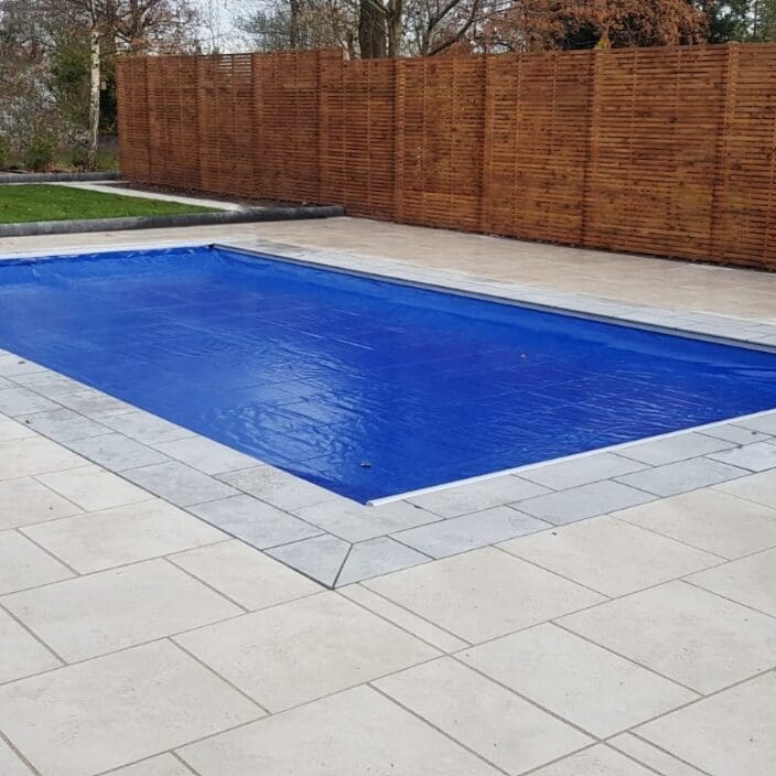 Rectangular in-ground pool covered by a taut blue cover, set within pale stone coping, surrounded by light tiled patio and tall wooden slatted fence with lawn and bare trees beyond.