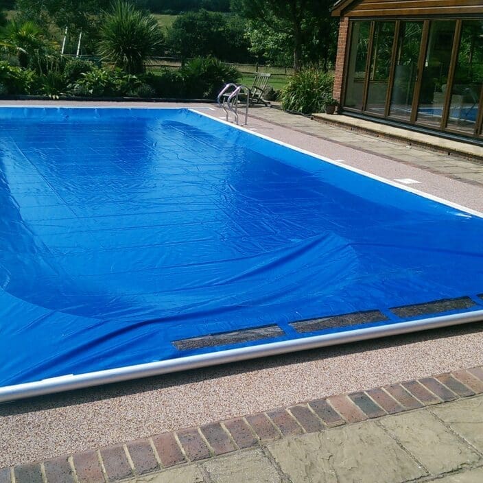 Rectangular swimming pool covered by a blue solar cover, slightly wrinkled and secured at one end, set beside a brick house with glass doors and a surrounding patio and garden.