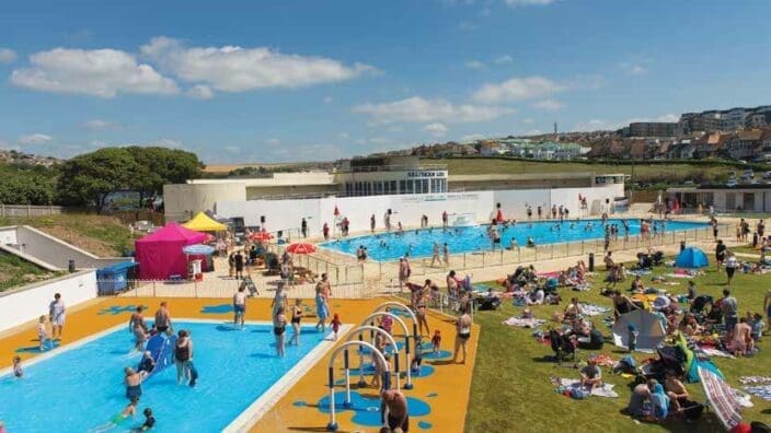 Outdoor lido SALTDEAN LIDO with families swimming and children playing in splash pools while others sunbathe on a grassy terrace under a clear blue sky beside a seaside town.