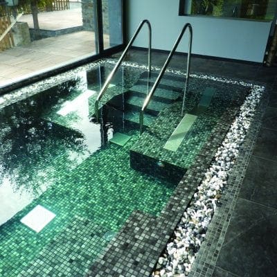 A small indoor mosaic-tiled plunge pool with metal handrails, stepped seating descends into clear green water, light reflecting on tiles, pebble border and slate floor beside large glass windows.