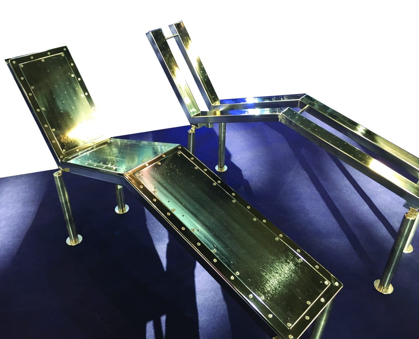 Polished brass-and-black metal lounge chairs extend and recline on a deep blue carpet, panels riveted and reflecting bright light, casting long angular shadows across the floor.