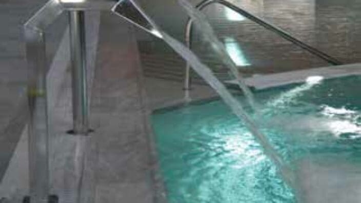 Stainless steel spouts pour two smooth streams into a turquoise indoor pool beside marble decking and curved metal handrails, producing ripples and bubbles under subdued overhead lighting.