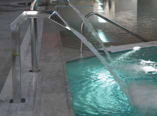 Stainless steel spouts pour two smooth streams into a turquoise indoor pool beside marble decking and curved metal handrails, producing ripples and bubbles under subdued overhead lighting.