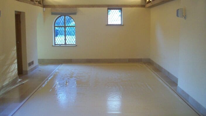 Rectangular indoor pool covered by a taut beige protective sheet, recessed into tiled floor; room features cream walls, exposed wooden roof beams, two diamond‑pane windows and soft overhead lighting.