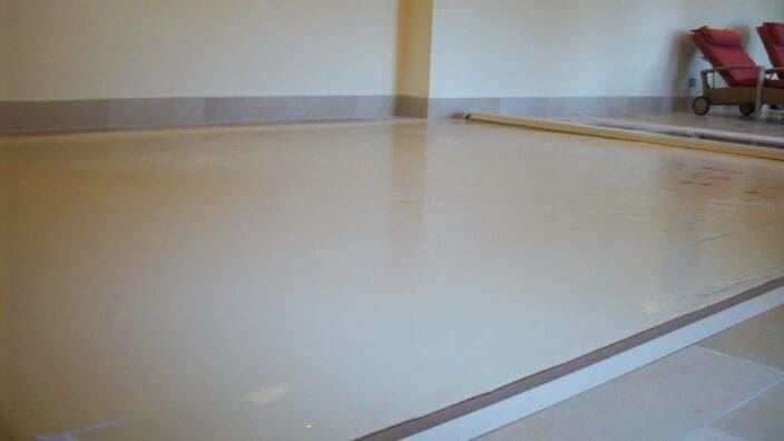 A large rectangular indoor pool covered with taut beige vinyl, recessed into a tiled floor; two red padded lounge chairs and a small leaded window sit against cream-colored walls.