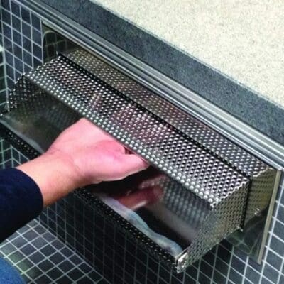 A stainless-steel perforated wall-mounted dispenser, a hand reaching under its lip to retrieve a towel or paper, installed in mosaic-tiled wall beneath a countertop edge.