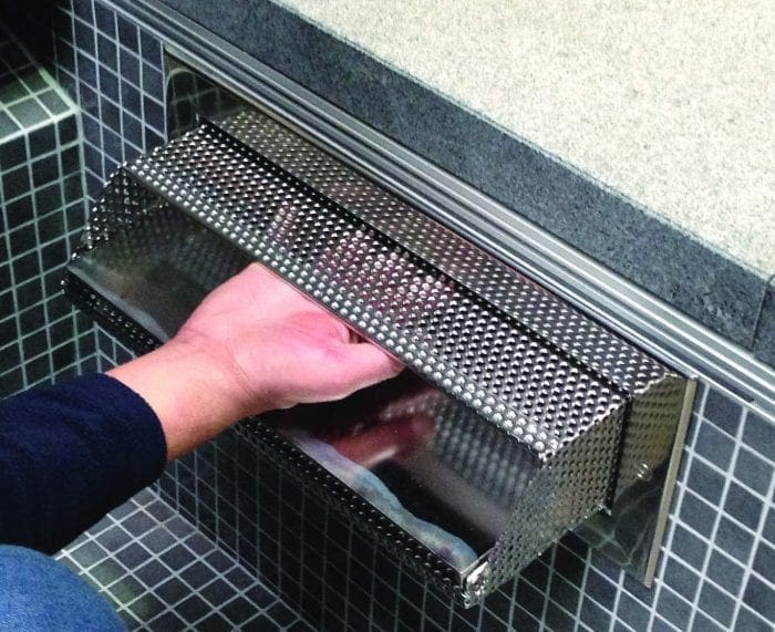 A stainless-steel perforated wall-mounted dispenser, a hand reaching under its lip to retrieve a towel or paper, installed in mosaic-tiled wall beneath a countertop edge.