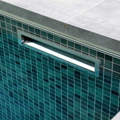 Rectangular recessed pool light glowing brightly in a teal mosaic-tiled wall, mounted just below gray concrete coping at the pool edge, reflecting subtle underwater light across tiles.