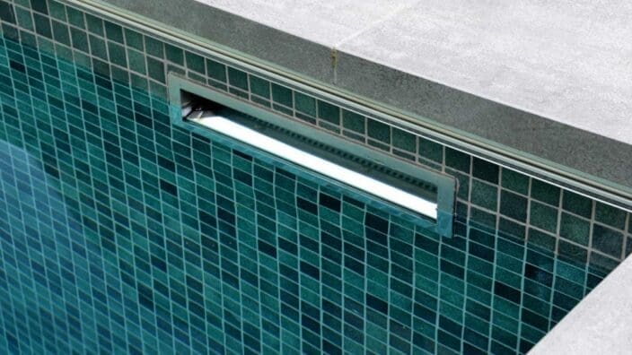 Rectangular recessed pool light glowing brightly in a teal mosaic-tiled wall, mounted just below gray concrete coping at the pool edge, reflecting subtle underwater light across tiles.