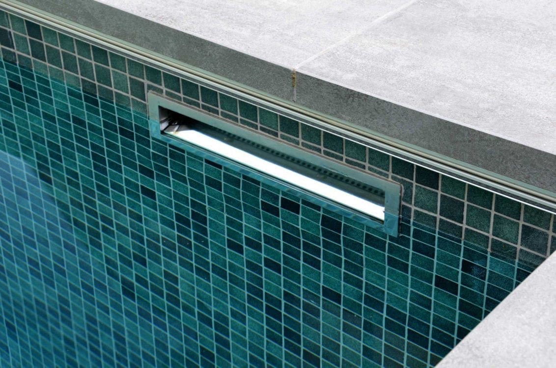 Rectangular recessed pool light glowing brightly in a teal mosaic-tiled wall, mounted just below gray concrete coping at the pool edge, reflecting subtle underwater light across tiles.