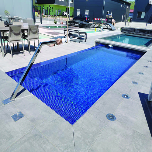 Rectangular blue-tiled plunge pool glistening under sunlight, with stainless handrail and grey stone deck, surrounded by patio furniture and neighboring pools in a modern outdoor display area, text 34