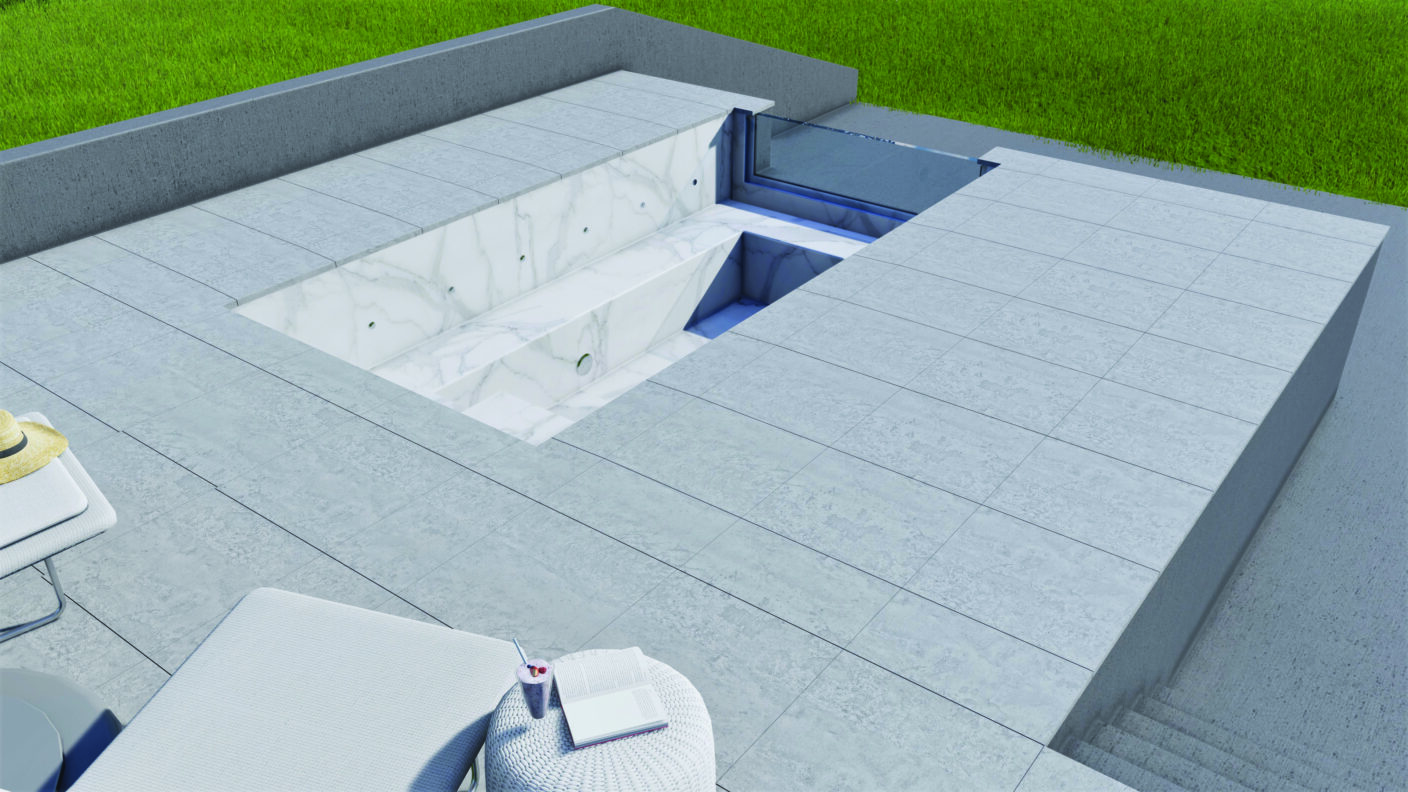 Sunken marble spa sits empty, revealing angled seats and plumbing holes; broad gray tiled terrace surrounds it with a lounge chair, table holding a hat, drink and book, grass beyond.