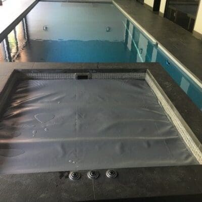 A square spa hot tub covered by a grey tarp sits empty in the foreground while a long indoor swimming pool stretches behind, surrounded by tiled deck and tall windows.