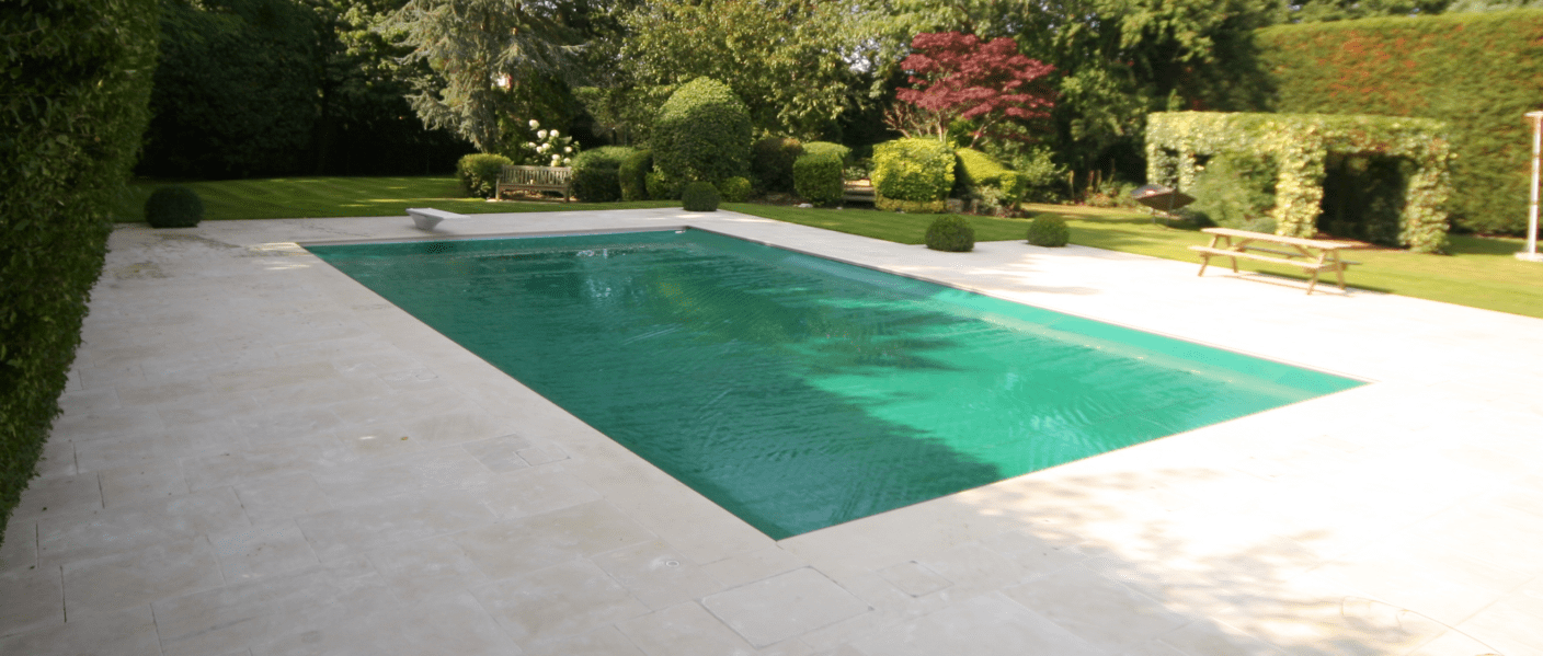 Rectangular swimming pool reflects sunlight, water rippling gently; surrounded by pale stone deck, trimmed hedges, manicured lawn, wooden bench and picnic table in a landscaped garden.