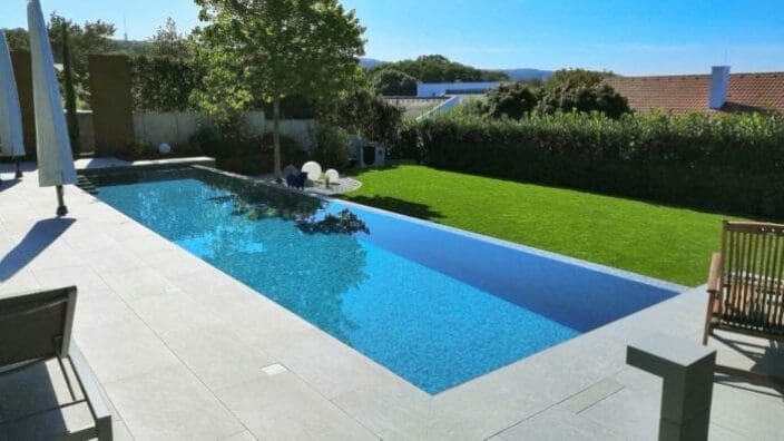 A rectangular infinity pool reflects a tree and blue sky, bordered by pale tiled terrace with lounge chairs and umbrellas, adjacent to a manicured green lawn and hedge-backed suburban houses.