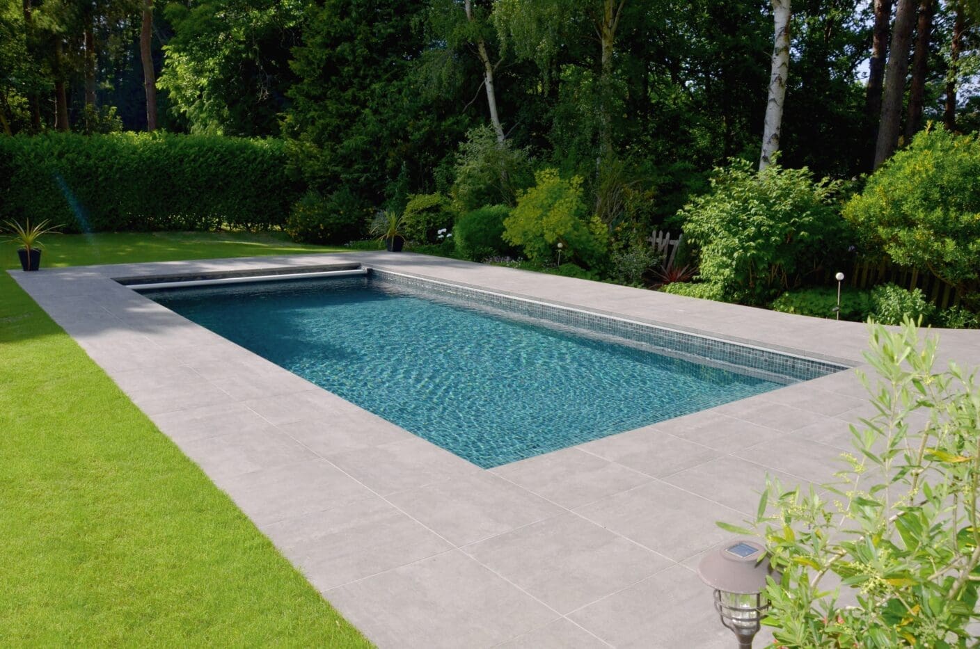Rectangular in-ground swimming pool ripples with clear blue water, bordered by wide gray tiles and neat green lawn, flanked by potted plants, shrubs and tall trees in a backyard.