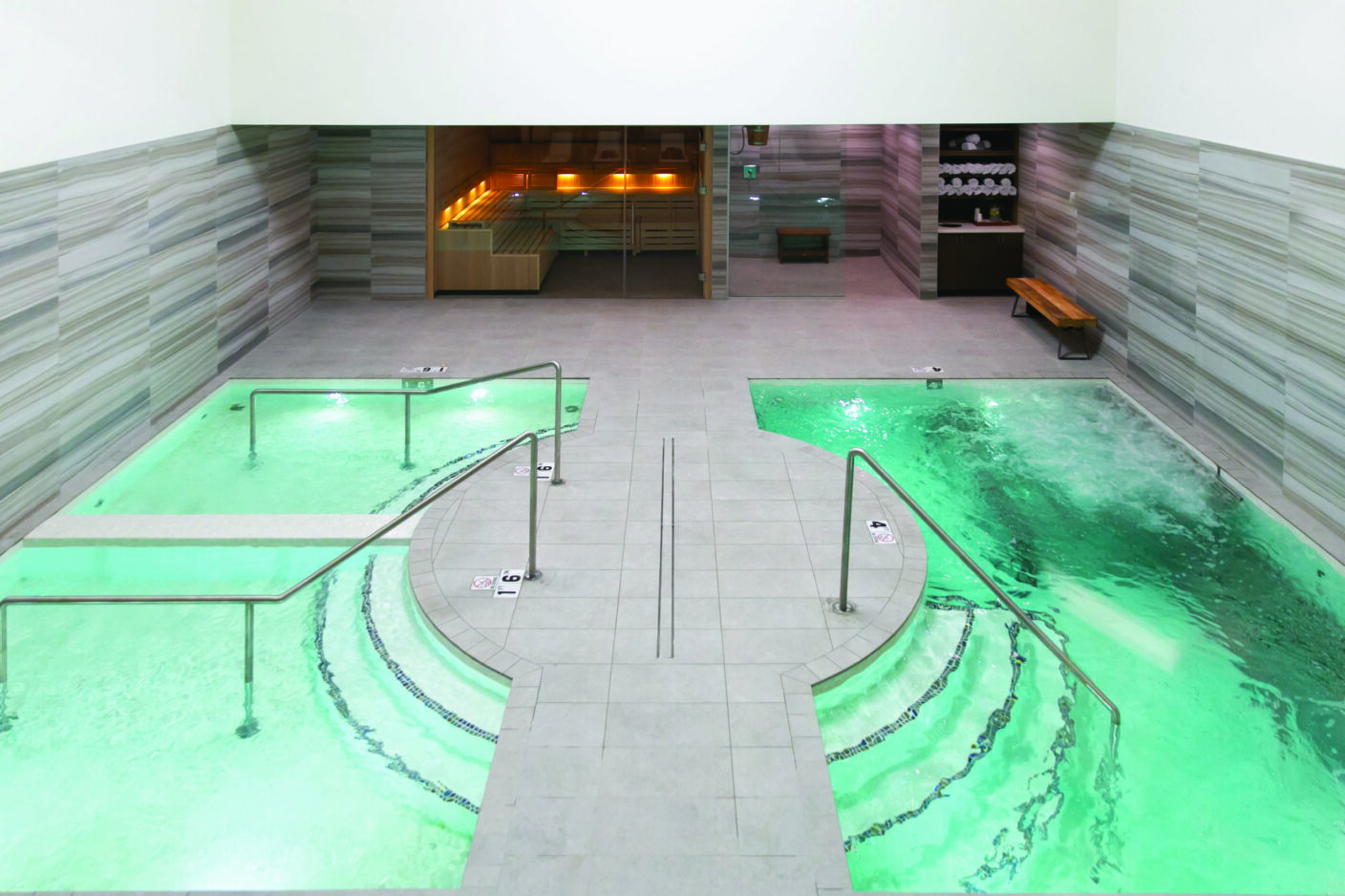 Two adjacent spa pools glowing green, bubbling and gently circulating, flanking a tiled central walkway with metal handrails; background shows glass-doored sauna and towel shelf. Text visible: 1.6, 4.