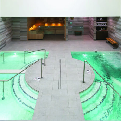 Two adjacent spa pools glowing green, bubbling and gently circulating, flanking a tiled central walkway with metal handrails; background shows glass-doored sauna and towel shelf. Text visible: 1.6, 4.