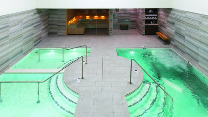 Two adjacent spa pools glowing green, bubbling and gently circulating, flanking a tiled central walkway with metal handrails; background shows glass-doored sauna and towel shelf. Text visible: 1.6, 4.